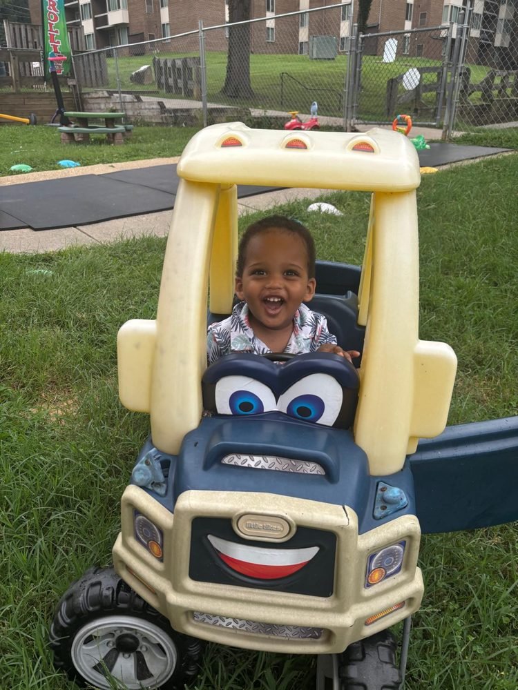 A toddler driving a toy truck in the EduCastle yard